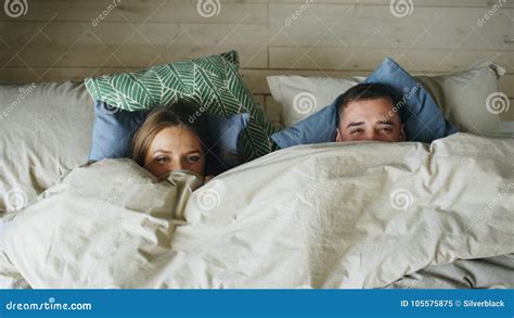 Top View Of Smiling Couple Having Fun In Bed Hiding Under Blanket And Looking Into Camera Stock