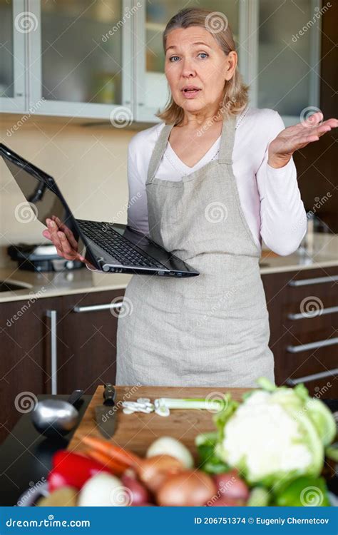 Mature Woman In The Kitchen Preparing Food And Holding Laptop Stock