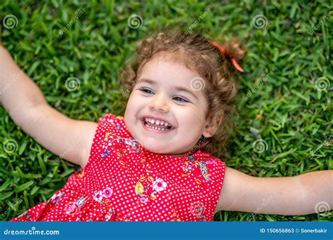 Happy Smiling Little Toddler Girl Laying On Grass In Park With Red