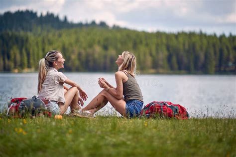 Young Female Friends Are Sitting On The Ground Beside The Lake While