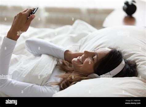 Woman Lying In Bed Listening Music Using Phone And Headphones Stock