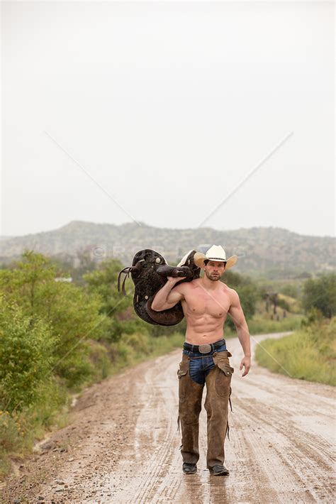 Muscular Shirtless Cowboy With A Saddle Over His Shoulder On A Dirt