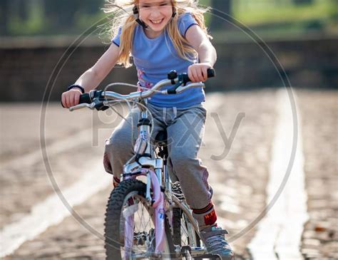 Image Of A Very Happy Young Blonde Girl Rides A Bike Along A Cobbled Path Towards Camera In
