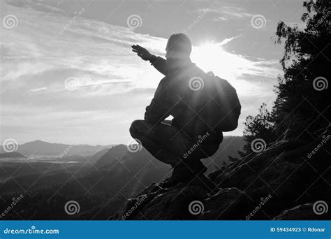 Hiker With Sporty Backpack Sit On Rocky Cliff Edge And Watching Into Misty Valley Bellow Sunny