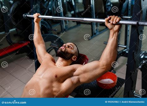 An Attractive Man With A Muscular Body Performs A Bench Press Using A Barbell On A Blurred Dark