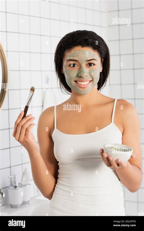 Smiling African American Woman With Mud Mask On Face Holding Cosmetic Brush And Bowl In Bathroom