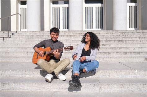 A Young Latin Couple Playing The Guitar And Laughing Sitting On Stairs