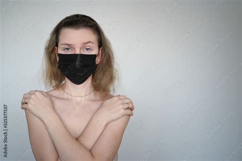 A Naked Girl In A Black Medical Mask Crossed Her Arms Over Her Chest Against A White Background