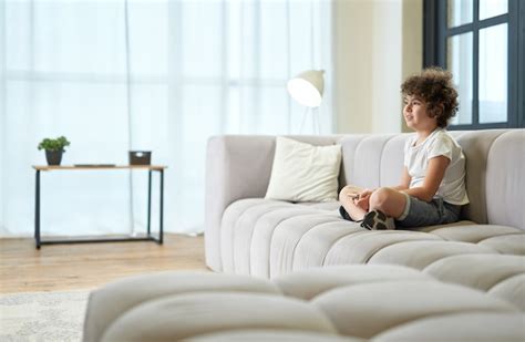 Adorable Niño Latino Sentado En Un Sofá Con Teléfono Inteligente Y Viendo La Televisión Mientras