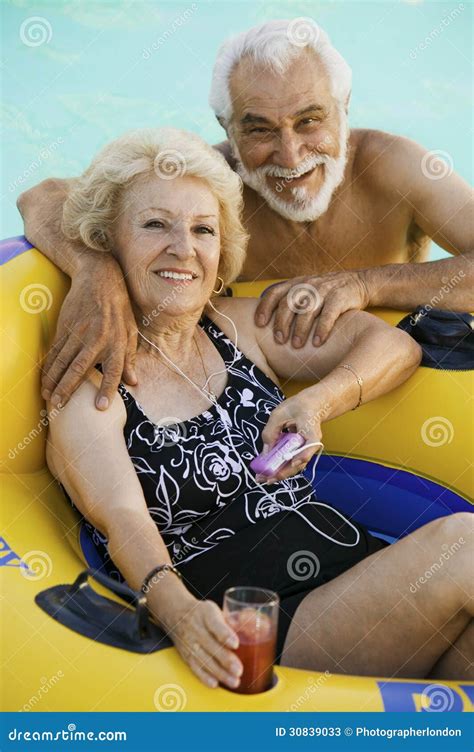 Senior Couple In Swimming Pool Woman Lying On Inflatable Raft Holding Drink Listening To