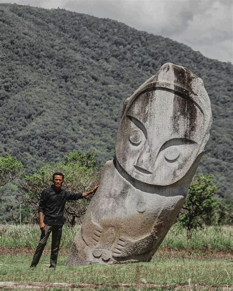 A Man Standing In Front Of A Large Rock With A Face Carved Into The Side