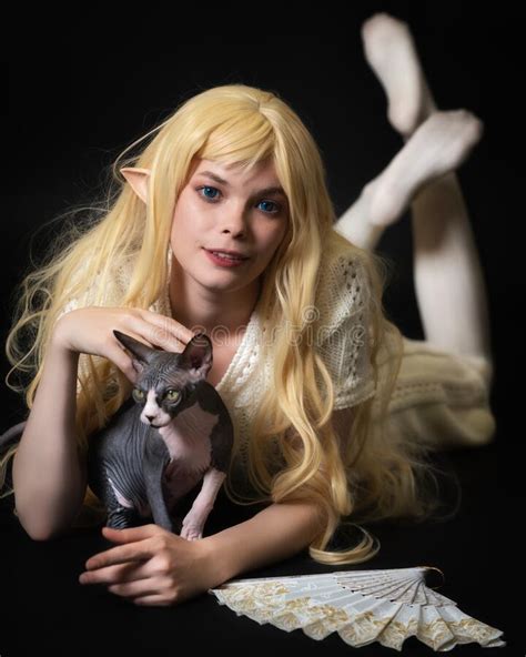 playful blue eyed blonde female elf cosplayer lying down on stomach and stroking sphinx kitten