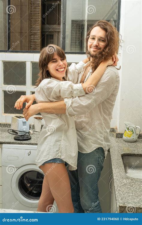 Young Attractive Couple In Love Hugging In The Kitchen While They Are Doing The Dishes Stock