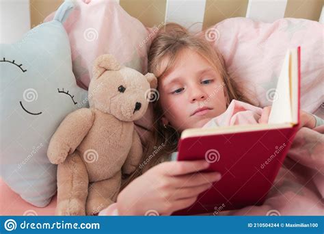 little blonde girl readingbook girl lying on white bed and reading book with her toy bear