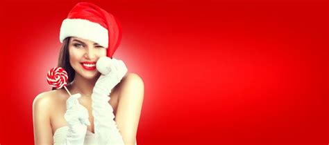 زن کریسمس دختر مدل زیبایی در کلاه بابا نوئل با لبهای قرمز دندانهای سفید و آب نبات شیرین خوری گل
