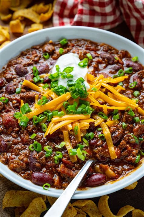 A White Bowl Filled With Chili And Cheese On Top Of Tortilla Chips Next To A Red Checkered Napkin