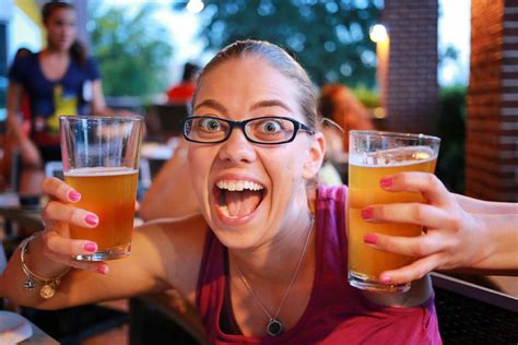 A Woman With Glasses Holding Two Beers In Front Of Her Face And Smiling At The Camera