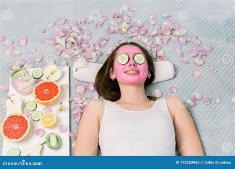 Beautiful Young Woman Is Getting Facial Clay Mask At Spa Lying With Cucumbers On Eyes Stock