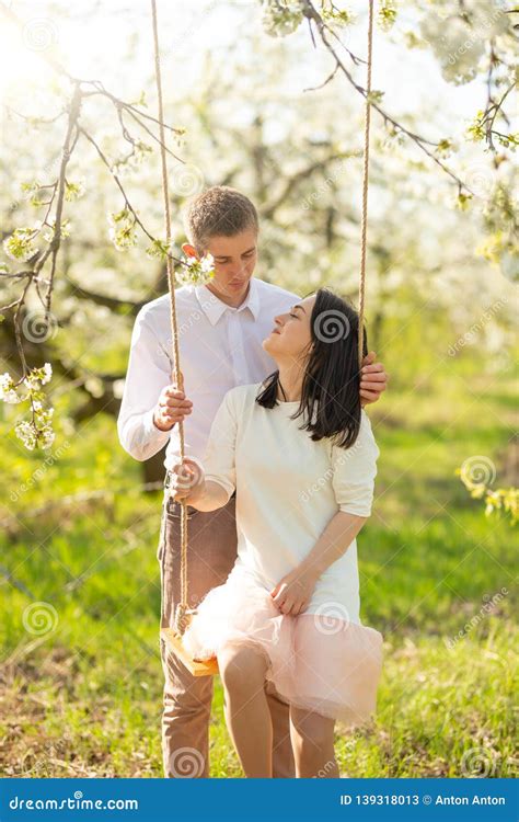 Young Married Couple On A Swing In A Flowered Garden Or Park Warmly