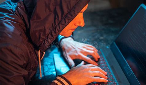 Hooded Hacker Is Typing On A Laptop Keyboard In A Dark Room Under A