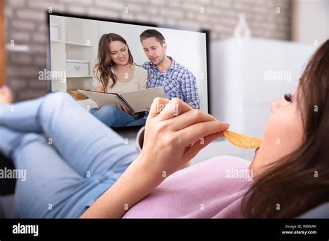 Woman Relaxing On Sofa Eating Potato Chips While Watching Television At
