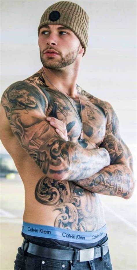 A Man With Tattoos On His Chest And Arms Is Standing In Front Of The Camera