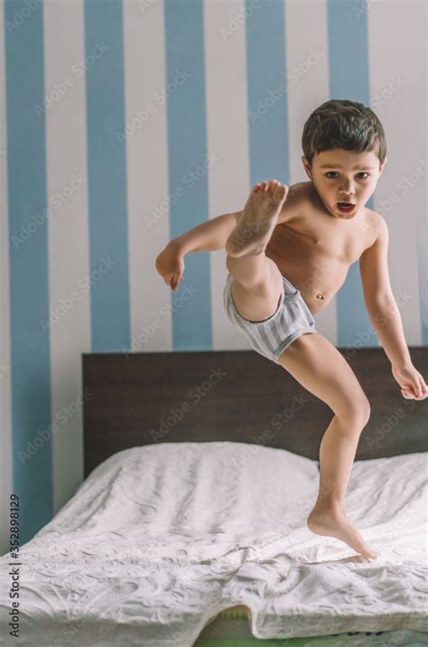 Cute Boy Having Fun While Jumping And Kicking With Leg On Bed Stock