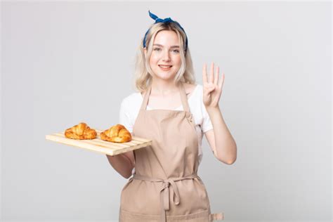 A Woman In An Apron Holding A Tray With Croissants On It And Waving