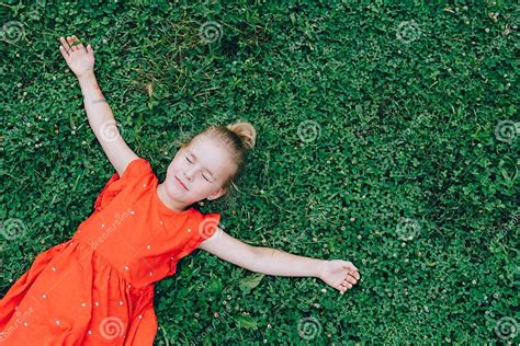 Smiling Little Cute Girl With Eyes Closed In Light Red Dress Lie On Green Grass Lawn Spreading