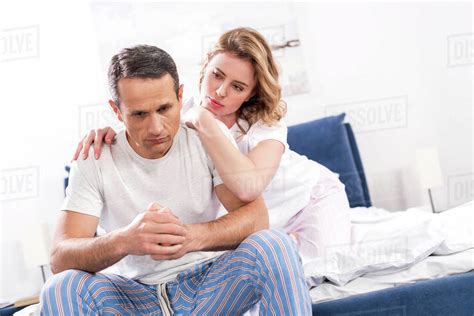 Woman Hugging Sad Husband That Sitting On Bed At Home Stock Photo