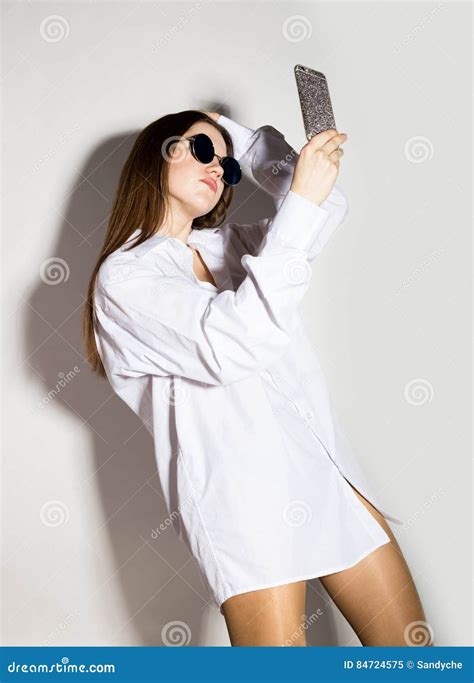 naked girl in a man`s white shirt and sunglasses holding phone doing selfie stock image