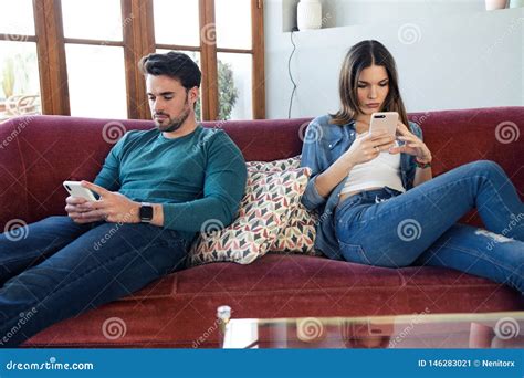 Angry Young Couple Ignoring Each Other Using Phone After An Argument While Sitting On Sofa At