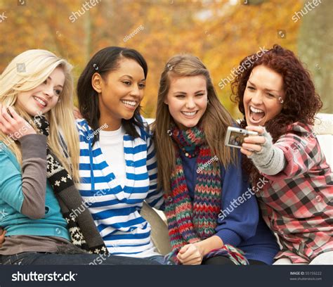 Group Of Four Teenage Girls Taking Picture With Camera Sitting On Bench In Autumn Park Stock