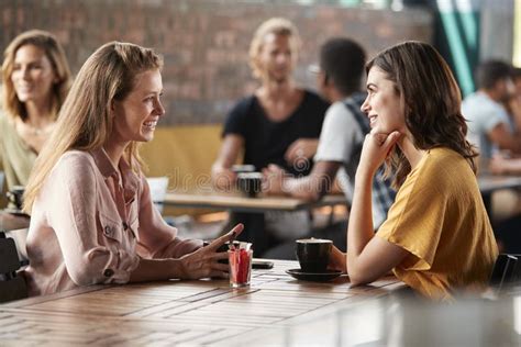 Two Female Friends Sitting At Table In Coffee Shop And Talking Stock