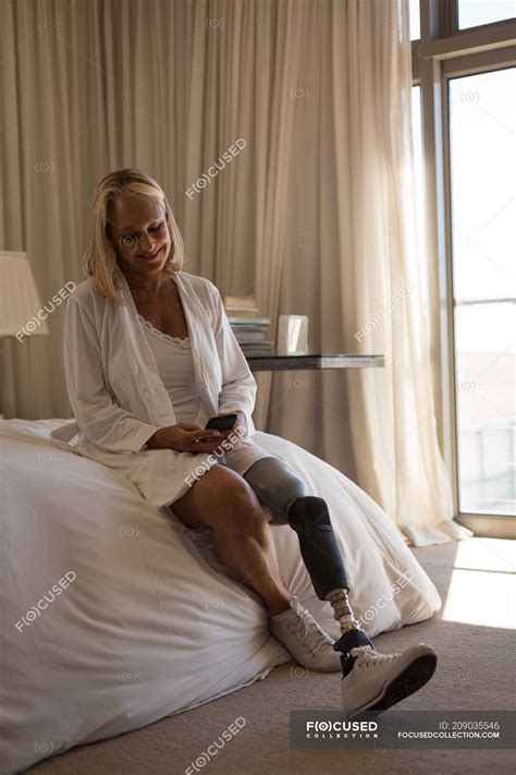 Mature Woman With Prosthetic Leg Using Mobile Phone In Bedroom At Home