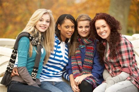 Group Of Four Teenage Girls Sitting On Bench In Autumn Park Stock