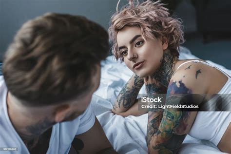 Young Tattooed Couple Looking At Each Other And Lying On Bed In Bedroom