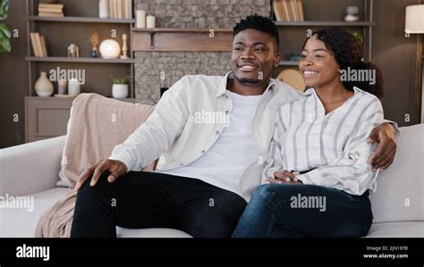 happy newlywed couple married african man and woman sitting at home sofa watching tv talking