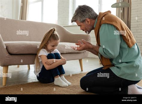 Strict Father Scolding Unhappy Daughter Sitting On Floor At Home Stock