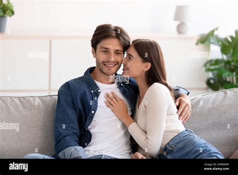 Cheerful Millennial European Woman Kissing Happy Guy On Cheek Sitting On Couch Look At Camera