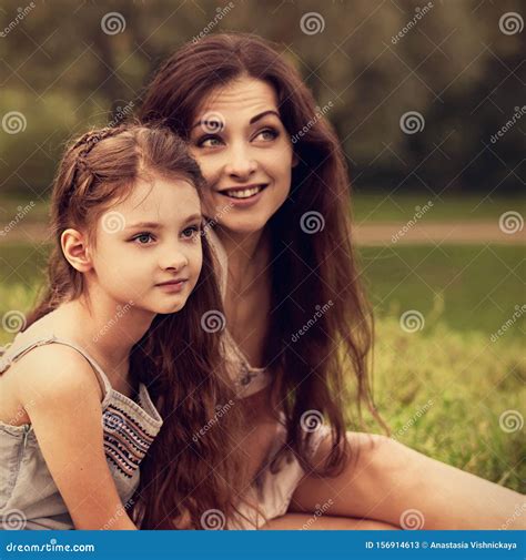 Beautiful Excited Mother Sitting With Her Cute Serious Daughter On Summer Green Grass Background