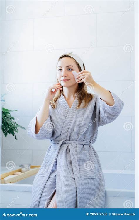 Young Woman In Bathrobe Looking In Mirror And Making Face Massage With Gua Sha Roller In