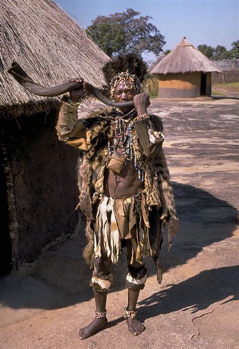 A Man In Native Clothing Standing Next To A Hut