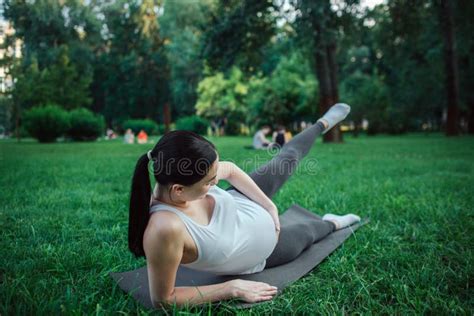 Serious And Concentrated Young Pregnant Woman Exercising On Yoga Mate In Park She Hold One Leg