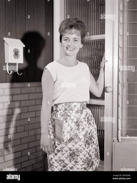 1960s Smiling Housewife Wearing Apron Standing With Front Door Ajar