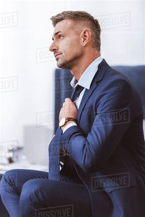Side View Of Handsome Man In Suit Sitting And Wearing Necktie At Home
