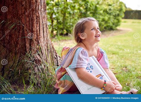 Smiling Young Girl Sitting Under Tree In Garden Reading Story Book