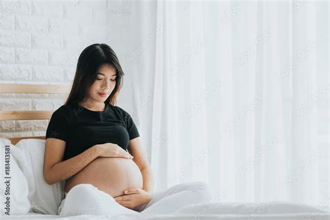 Happy Asian Pregnant Woman Touching Her Belly With Care In Bedroom She