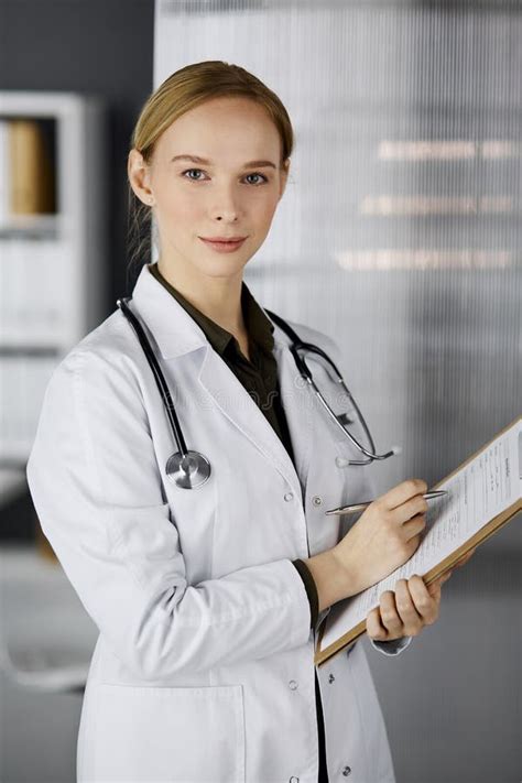 Friendly Smiling Female Doctor Using Clipboard In Clinic Portrait Of