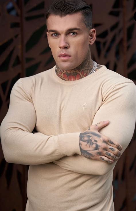 A Man With Tattoos Standing In Front Of A Wooden Wall Wearing A Tan